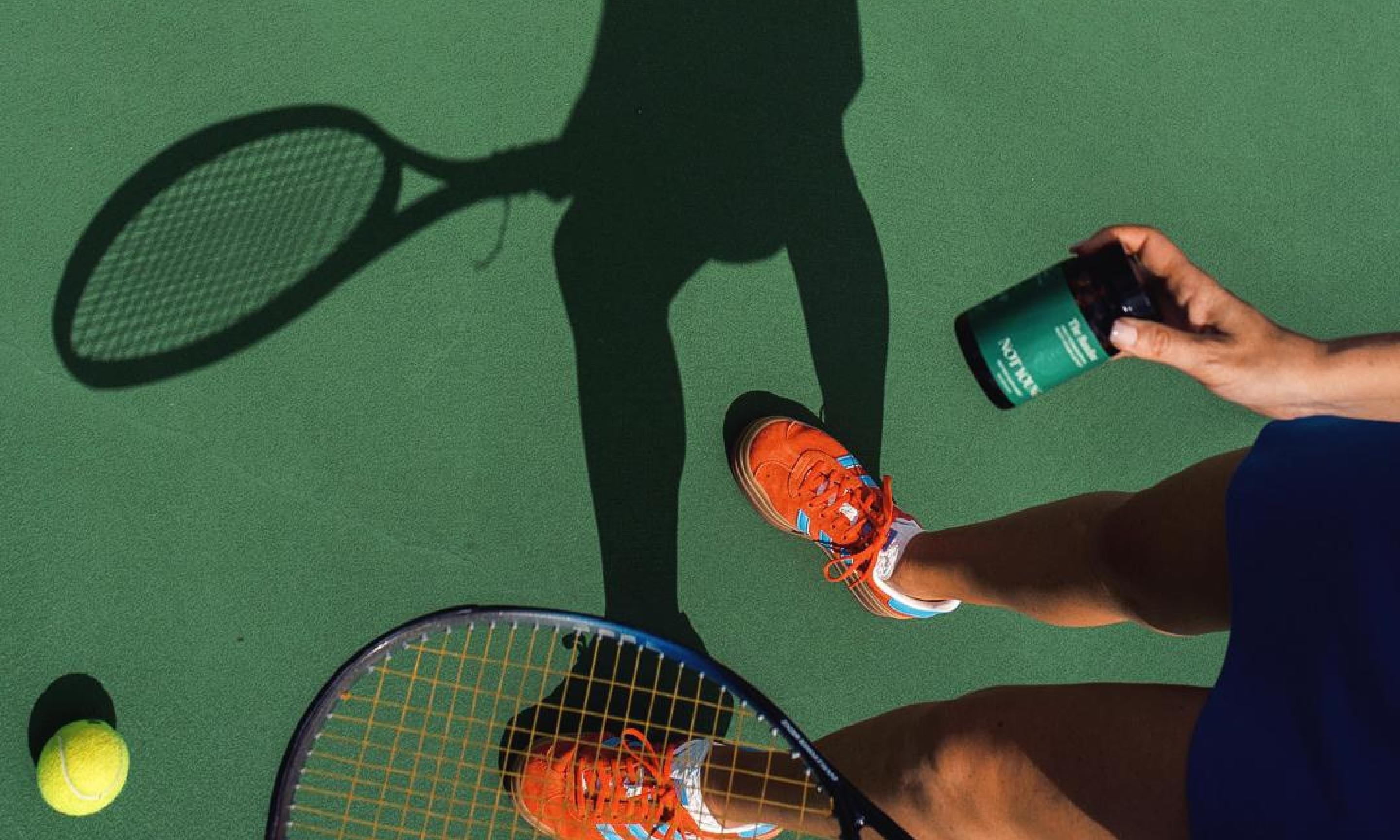 A photography of a tenis court background with The Basline being held by a woman in one hand with a tennis racket in the other